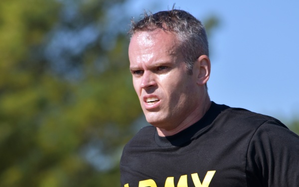 A male individual is running outdoors while wearing a black U.S. Army physical training t-shirt with yellow lettering. His expression is intense and focused, and visible perspiration suggests sustained physical exertion. The background features blurred green trees and a clear blue sky, indicating an outdoor daytime setting.
