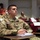 A group of U.S. Air Force personnel are seated in a classroom or auditorium-style setting, attending a briefing or training session. The foreground subject, wearing OCP (Operational Camouflage Pattern) uniform with 'U.S. AIR FORCE' tape and a name tape reading 'STEIGER,' holds a pen over a notebook. Social distancing measures are visible, indicated by 'DO NOT SIT HERE' signs on certain seats.
