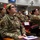 A group of military personnel in OCP (Operational Camouflage Pattern) uniforms are seated in an auditorium-style briefing room, attentively watching a presentation. The foreground shows a female airman wearing glasses with a notebook and pens on her desk armrest. A 'DO NOT SIT HERE' sign is visible on one of the seats, suggesting COVID-19 social distancing protocols.