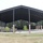 An open-sided metal pavilion structure with a dark roof supported by brick-based steel columns, housing what appears to be military vehicle displays on a concrete pad. The facility appears to be an outdoor military museum or memorial exhibit area. Young trees have been recently planted around the perimeter of the structure.
