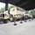 Multiple tan/desert-camouflage military vehicles are displayed under a covered open-air pavilion structure. The vehicles are cordoned off with black stanchion posts connected by yellow-and-black caution chains, suggesting a static display or museum-style exhibit. The setting appears to be a military installation or heritage display area surrounded by pine trees.
