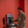 A military service member in MultiCam/OCP camouflage uniform is filling a dark green water bottle at an Elkay EZH2O bottle-filling station mounted on a red wall. The individual appears to be in an indoor facility, likely on a military installation. A US Air Force patch is visible on the left shoulder of the uniform.