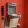 The image shows an Elkay EZH2O combination drinking fountain and bottle-filling station mounted on a terracotta/red wall. The unit is made of stainless steel and includes both a traditional drinking spout and an upper bottle-filling module with a digital counter display. An electrical outlet with cables is visible at the base of the unit.