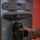 A person in military camouflage uniform is filling a dark olive green insulated water bottle at an Elkay EZH2O bottle-filling station. The stainless steel hydration station features a filter status indicator and a bottle counter display. The water bottle appears to have a logo or text reading 'QATAR' on its side.