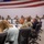 A formal military briefing or conference is taking place in a large meeting room with multiple rows of participants seated at long tables. The majority of attendees are wearing U.S. Army combat uniforms (ACU/OCP pattern), with a few civilians also present. A large American flag is prominently displayed on the wall at the front of the room.