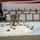 A military officer in camouflage uniform is positioning unit markers on a large floor map during what appears to be a command post exercise or war game. An audience of approximately 20 uniformed military personnel observes from elevated seating in the background. The setting appears to be a large indoor facility, possibly a hangar, decorated with an American flag.