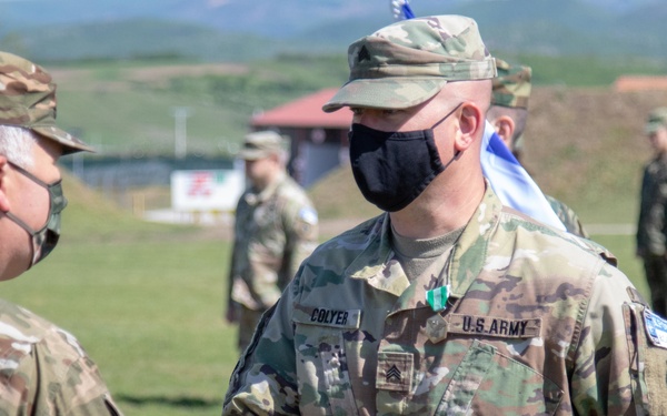 Two U.S. Army soldiers are shaking hands during what appears to be a military ceremony or award presentation outdoors. The soldier on the right, identified by name tape as 'COLYER' with the rank of Sergeant, is receiving what appears to be a medal or commendation. Both soldiers are wearing black face masks and MultiCam (OCP) uniforms, with additional military personnel visible in the background.