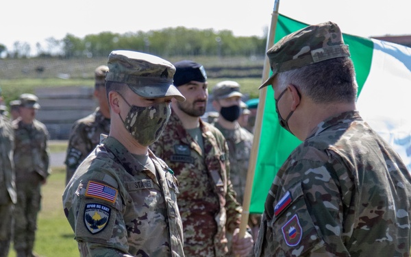 A military ceremony showing two soldiers shaking hands in the foreground, with one wearing a US Army patch and an American flag on their sleeve, and the other displaying what appears to be a Slovenian flag patch. An Italian flag is visible being carried in the background. Multiple military personnel in various camouflage uniforms are assembled in formation, several wearing face masks consistent with COVID-era protocols.