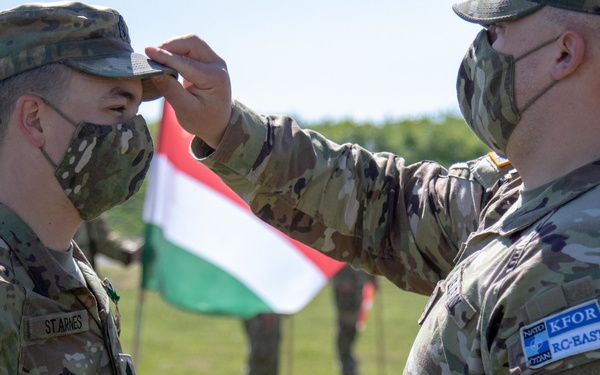 Two U.S. Army soldiers in OCP (Operational Camouflage Pattern) uniforms are engaged in what appears to be a military ceremony, with one soldier adjusting or pinning something to the other's cap. Both wear COVID-19 face masks and display KFOR RC-East patches, indicating deployment with NATO's Kosovo Force. A Hungarian flag is visible in the background along with additional personnel.