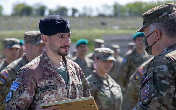 A military ceremony is taking place outdoors, where a soldier wearing Italian Army ('ESERCITO') camouflage uniform and black beret is receiving or exchanging a framed item with another soldier wearing a face mask and a Slovenian flag patch. Multiple soldiers in various uniforms are assembled in formation in the background, many wearing teal/green berets suggesting a multinational military exercise or event. The setting appears to be a training ground or military field area.