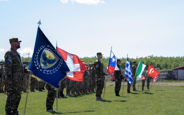 A multinational military ceremony is taking place on an open grass field, with soldiers in camouflage uniforms standing in formation. Multiple national flags are being carried by flag bearers in a line, suggesting a joint military exercise, parade, or official ceremony. The lead soldier prominently displays a blue organizational flag bearing an eagle emblem and laurel wreath.