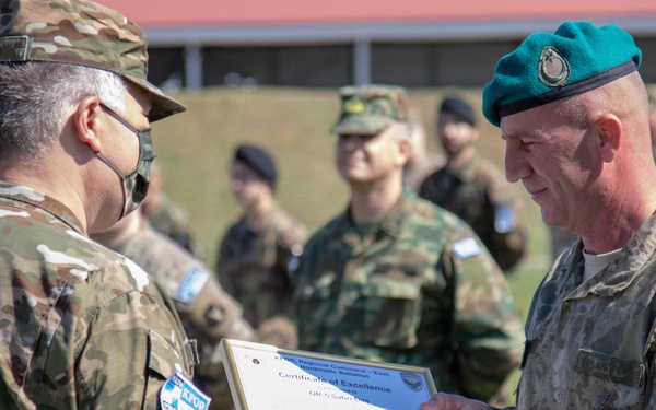 A military ceremony is taking place outdoors where a Turkish soldier wearing a green beret is presenting a 'Certificate of Excellence' to another soldier wearing a face mask. The recipient appears to be from a multinational unit based on the patches visible on his uniform. Other military personnel in various uniforms are visible in the background observing the ceremony.
