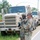 Two U.S. Army soldiers in Operational Camouflage Pattern (OCP) uniforms stand alongside a road next to a convoy of military trucks. One soldier wearing a combat helmet appears to be speaking on a radio while the other, wearing a patrol cap, listens with arms crossed. The setting appears to be a training environment based on the lush vegetation and road markings.