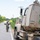 A U.S. Army soldier in full combat gear is directing or guiding a large military tactical truck (tan/desert-colored) that is pulled over on the shoulder of a rural road. The truck is marked with unit identifiers '642RSG' and '828BN', indicating it belongs to the 642nd Regional Support Group, 828th Battalion. The scene appears to be a convoy operation or training exercise on a two-lane road lined with trees and utility poles.