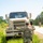 A soldier in OCP (Operational Camouflage Pattern) uniform, identified by nametape as 'DEAN', is directing a military truck (appears to be an LMTV or similar tactical vehicle) that has pulled off the road onto a grassy shoulder. The soldier has both hands raised in a halting or guiding gesture while wearing black tactical gloves. A second soldier is visible in the background on the road.