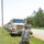 Two U.S. Army soldiers in full combat gear are walking alongside a convoy of tan/beige military trucks (likely LMTV or HEMTT variants) parked on a grassy roadside. The foreground soldier appears to be directing or inspecting the convoy, with one arm extended toward the lead vehicle. The setting appears to be a rural training area with trees and a paved road visible.