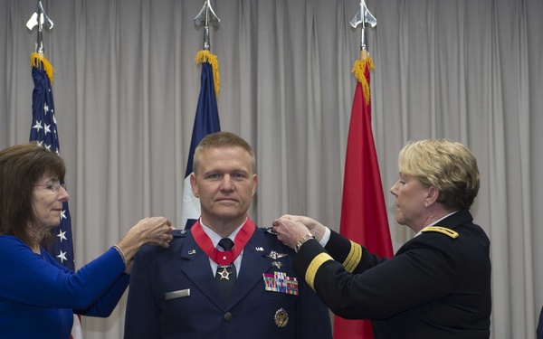 A military promotion or decoration ceremony is taking place in a formal indoor setting. A male U.S. Air Force officer in dress blues is having insignia or rank devices pinned to his shoulders simultaneously by a civilian woman on his left and a female U.S. Army general officer on his right. A female Air Force Master Sergeant stands in the background observing the ceremony.