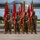 A color guard formation of U.S. Marines in MARPAT camouflage uniforms and black face masks stands at attention on an airfield tarmac, holding multiple red and gold unit guidons and flags. The Marines are positioned in front of a large military transport aircraft, likely a C-130 Hercules. The scene appears to be a formal military ceremony, possibly a change of command or unit activation event.