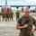A senior U.S. Marines officer, identified by the name tape 'CLARDY' and two-star general rank insignia, speaks into a microphone on a military flight line. Behind him, a formation of Marines in camouflage uniforms stands in front of a large military turboprop aircraft, with multiple military flags and colors visible. The setting appears to be a formal military ceremony or change of command event.