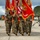 A U.S. Marine Corps color guard is marching in formation on what appears to be a military airfield or tarmac. The personnel are carrying multiple unit guidons and organizational flags with gold fringe, consistent with a formal military ceremony such as a change of command or activation/deactivation event. All personnel are wearing face masks, suggesting this was taken during the COVID-19 pandemic era.