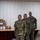 A group of U.S. Air Force personnel in OCP (Operational Camouflage Pattern) uniforms are standing at attention during what appears to be a formal military ceremony or formation. The setting appears to be an indoor facility, possibly a dining facility or multipurpose room, with a U.S. flag visible on the left side. At least one individual in the background is wearing a black face mask, suggesting this event occurred during the COVID-19 pandemic era.