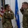 Two U.S. Air Force personnel in OCP (Operational Camouflage Pattern) uniforms are exchanging salutes during what appears to be a formal military ceremony. Both individuals are facing each other in a saluting position, with American and Air Force flags visible in the background. A third person is partially visible on the left holding what appears to be a blue presentation folder or award case.