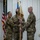 A military change of command ceremony is taking place indoors, with two service members in OCP (Operational Camouflage Pattern) uniforms exchanging a unit guidon. The guidon appears to read '380 EMS' or similar unit designation. A US flag and what appears to be an Air Force flag are visible in the background.