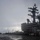 USS Ronald Reagan Conducts Flight Operations in the South China Sea The image shows the island superstructure of a U.S. Navy aircraft carrier bearing the hull number 76, identifiable as USS Ronald Reagan (CVN-76). The flight deck is wet, suggesting recent rain or overcast maritime conditions, and is populated with personnel and parked aircraft. The scene appears to be during flight deck operations or a stand-down period at sea.