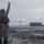 USS Ronald Reagan Conducts Flight Operations in the South China Sea A naval flight deck crew member wearing a red safety vest and helmet raises one hand while signaling on a wet aircraft carrier flight deck. A military jet aircraft, appearing to be an F-35 or similar stealth fighter, is on approach for landing in the background. Additional deck crew members are visible along the right side of the deck near what appears to be a jet blast deflector.