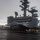 USS Ronald Reagan Conducts Flight Operations in the South China Sea A carrier-based jet aircraft, appearing to be an F/A-18 Hornet or Super Hornet, is positioned on the wet flight deck of a U.S. Navy aircraft carrier. The island superstructure is visible in the background, displaying the hull number '76', identifying this as the USS Ronald Reagan (CVN-76). The overcast, rainy conditions suggest flight operations during adverse weather.