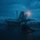 USS Ronald Reagan Conducts Flight Operations in the South China Sea A military jet aircraft, consistent with an F/A-18 Hornet or Super Hornet, is positioned on the flight deck of an aircraft carrier during twilight or pre-dawn conditions. A flight deck crew member is visible in a crouching or launch-signal posture in the foreground, suggesting an imminent catapult launch. The wet deck surface and overcast sky indicate adverse weather conditions during flight operations.