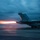 USS Ronald Reagan Conducts Flight Operations in the South China Sea A military jet aircraft, identifiable as an F/A-18 type, is captured during a catapult launch from what appears to be an aircraft carrier deck at dusk or dawn. The afterburners are visibly ignited, producing a bright orange flame plume extending behind the aircraft. The motion blur and low-light conditions create a dramatic, high-energy composition.