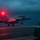 USS Ronald Reagan Conducts Flight Operations in the South China Sea A military jet aircraft, consistent with an F/A-18 Hornet or Super Hornet, is captured during a night or dusk catapult launch from what appears to be an aircraft carrier flight deck. The aircraft's afterburners are ignited and a bright red anti-collision beacon light is prominently visible. The wet reflective deck surface and overcast sky contribute to the dramatic low-light atmosphere.