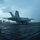 USS Ronald Reagan Conducts Flight Operations in the South China Sea A U.S. Navy carrier-based jet aircraft, marked with the number '201', is positioned on a wet aircraft carrier flight deck under overcast, low-light conditions. A flight deck crew member is visible in the foreground in a crouched launch signaling posture, while additional personnel are visible along the deck edge in the background. The scene appears to depict a catapult launch operation.