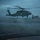 USS Ronald Reagan Conducts Flight Operations in the South China Sea A military helicopter, consistent with an MH-60 Seahawk, is hovering or landing on what appears to be a naval vessel's flight deck during severe weather conditions. Several deck crew members in flight deck gear are visible managing the operation in heavy rain and low visibility. A green navigation light is illuminated on the starboard side of the aircraft.