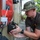 A military service member wearing digital camouflage uniform and boonie hat is closely inspecting or adjusting a mechanical component on the exterior of a military vehicle. The individual appears to be performing maintenance or equipment checks on what appears to be a latch or locking mechanism. The setting appears to be an outdoor military installation or staging area.