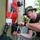 A military service member wearing a camouflage cover (hat) and digital camouflage trousers is working on a latch or locking mechanism on what appears to be a military vehicle or equipment container. The individual is using both hands to manipulate a black locking device, concentrating closely on the task. A red and white cross symbol is visible on the surface, suggesting possible medical or emergency equipment.