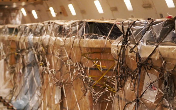 The image shows the interior of a cargo aircraft loaded with multiple pallets of freight secured with cargo nets and plastic wrapping. A USAID label is visible on one of the pallets, indicating humanitarian aid shipment. The cargo is arranged along a roller conveyor track system typical of wide-body freighter aircraft.