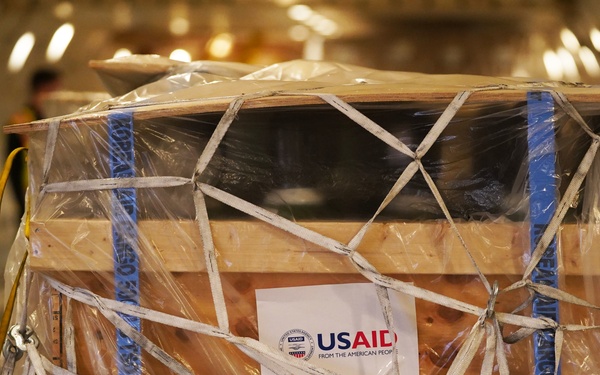 A large wooden crate secured with white cargo netting and blue strapping tape is loaded inside the cargo hold of an aircraft. The crate bears a USAID label reading 'From the American People,' indicating it contains humanitarian aid supplies. The cargo is wrapped in clear plastic sheeting for protection during transport.
