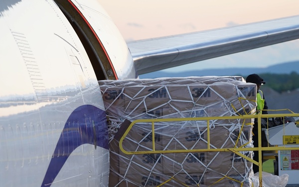 A cargo loading operation is underway at an airport, with a large pallet of 3M-branded boxes being loaded into the cargo hold of a commercial aircraft. The ground support equipment is branded 'Jet Aviation, A General Dynamics Company.' A worker in a high-visibility vest is visible on the loading platform overseeing the operation.