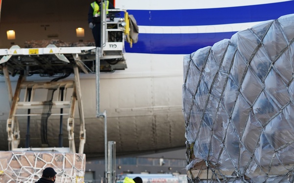The image shows a cargo loading operation at an airport, with workers in high-visibility vests handling palletized freight next to a large cargo aircraft. A USAID label is clearly visible on one of the shrink-wrapped pallets, indicating humanitarian or foreign aid shipment. Ground crew are actively positioning cargo on roller-equipped loading platforms near the open nose or cargo door of the aircraft.