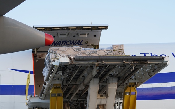 A cargo aircraft, partially visible with the text 'NATIONAL' and 'The World' on its fuselage, is being loaded or unloaded at an airport. A large cargo loader/scissor lift platform is positioned against the aircraft's main deck cargo door, with wrapped pallets visible on the platform. In the background, another aircraft with 'Omni Air Inter...' livery is partially visible.