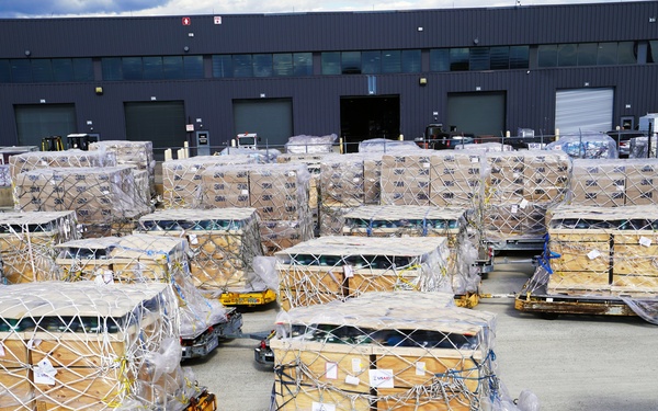 The image shows a large cargo staging area outside an industrial warehouse or freight terminal, with numerous palletized shipments loaded onto yellow airport-style cargo dollies. The cargo consists of wooden crates and cardboard boxes wrapped in plastic sheeting and secured with cargo nets and strapping. The scene suggests active air freight or logistics operations with goods awaiting loading or recently unloaded.