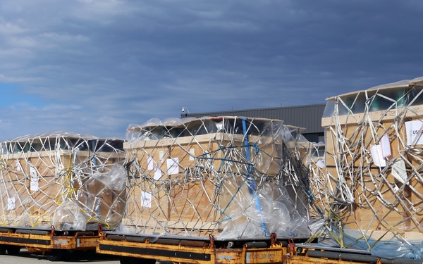 The image shows a row of large air cargo pallets staged on yellow ground support equipment (GSE) dollies on an airport tarmac or cargo apron. Each pallet contains wooden crate structures wrapped in clear plastic sheeting and secured with cargo nets. The setting appears to be an air freight facility, with a building visible in the background under a partly cloudy sky.