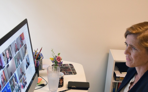 A woman with auburn hair in a braid is seated at a white desk, participating in a video conference call displayed on an Apple iMac monitor showing a grid of multiple participants. She is dressed in a dark blazer over a light-colored top and appears to be attentively watching the screen. The home office setting includes various desk items and printed documents visible in the foreground.