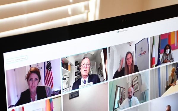 A computer monitor displays an active video conference call with approximately 12-15 participants visible in a grid layout, appearing to be an international diplomatic or governmental meeting. Multiple national flags and organizational logos are visible in participants' backgrounds, including USAID, the Canadian flag, the EU flag, the Japanese flag, and the American flag. The platform appears to be Cisco Webex, with a participant list panel visible on the right side of the screen.