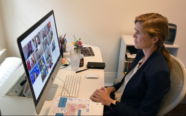 A woman with auburn hair in a braided ponytail sits at a white desk participating in a video conference call on an Apple iMac computer. She is dressed in a dark navy blazer and appears to be in a home office setting. The monitor displays a grid of multiple video call participants.
