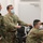 The image shows a group of military personnel from multiple nations gathered in what appears to be a command or operations room. At least three nationalities are identifiable through uniform patches, including German and American forces, all wearing blue surgical face masks. Personnel appear to be engaged in a briefing or monitoring activity at workstations.