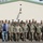 A large group of military personnel in camouflage uniforms pose for a formal group photograph in front of a building with a green roof. The group includes both male and female service members, with one civilian in a suit and tie visible on the left side. The setting appears to be a military installation, likely in Europe based on architectural style and the presence of what appear to be NATO allied uniforms.