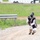 A person wearing a protective helmet/mask and loose white clothing is kneeling on a gravel path while holding what appears to be a paintball gun. The setting appears to be an outdoor recreational or training area with green grass, a guardrail, and vehicles visible in the blurred background. The attire and equipment suggest participation in a paintball or airsoft activity.
